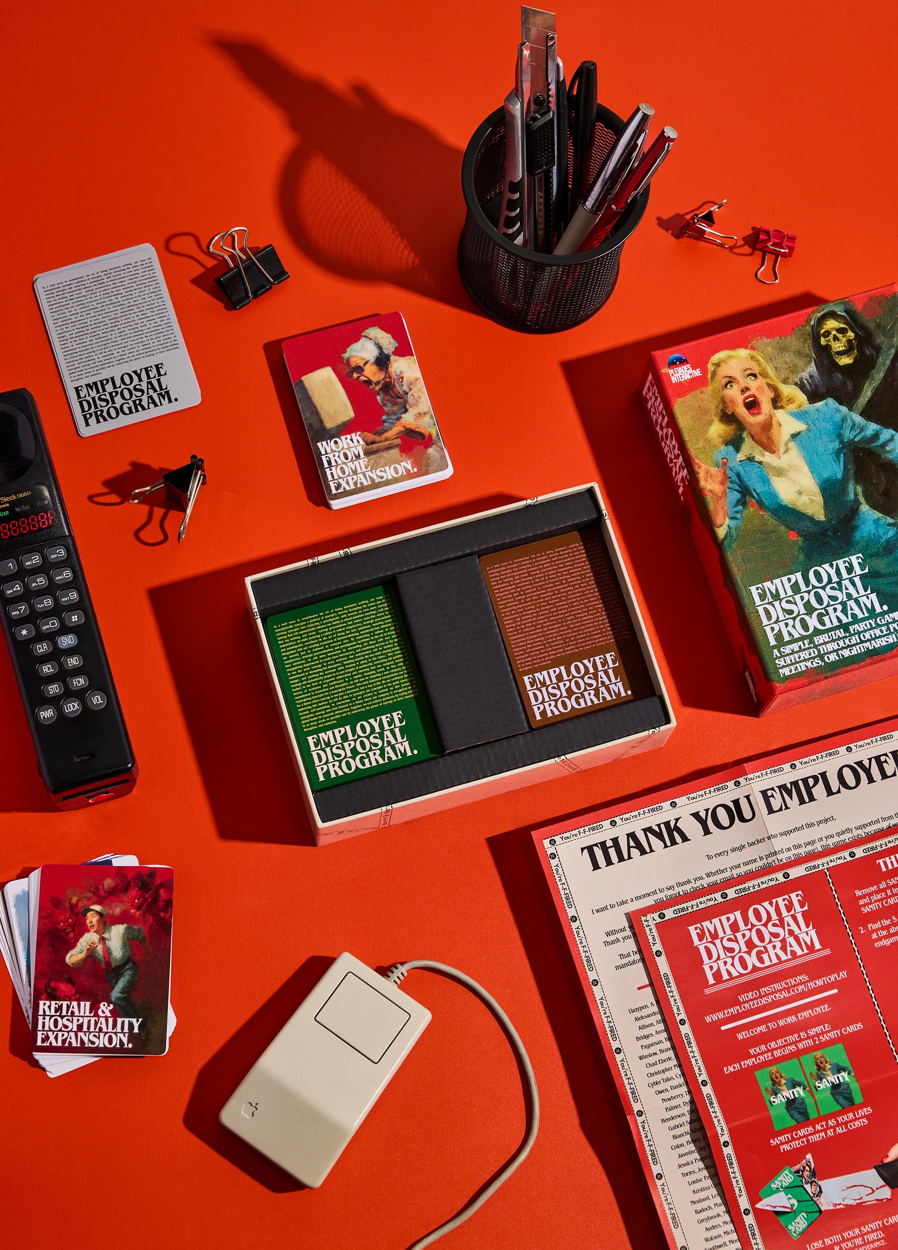 Employee Disposal Program Deluxe Edition box open on a red desk with expansion decks, bonus mini packs and rulebook